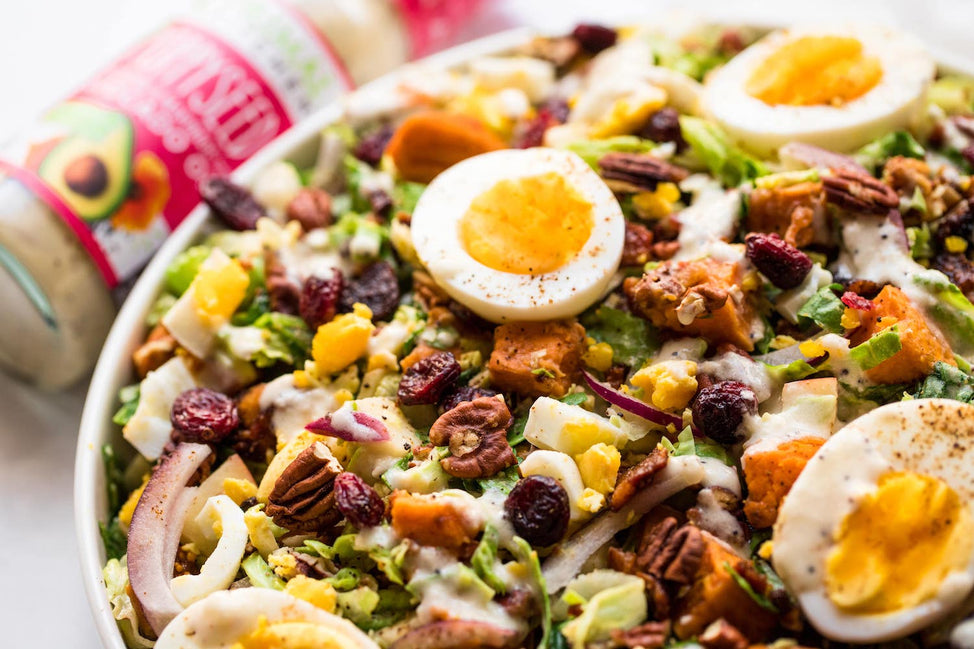 A white bowl filled with an autumn salad of bacon, cranberries, pecans and more, with a bottle of Primal Kitchen Poppyseed Dressing in the background.
