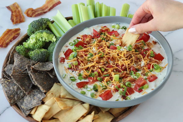 A hand dipping a chip into a bowl of Bacon Dip, with chips and veggies on the side.