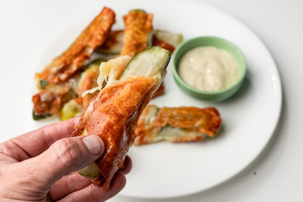 A hand holding a chickle over a plate of cheese-wrapped pickles with a container of Primal Kitchen Ranch for dipping.
