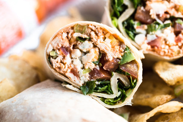 Closeup of a Buffalo chicken wrap made with Primal Kitchen Buffalo Sauce, cut in half. More wraps and potato chips on the sides.