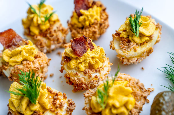 Closeup of fried pickle deviled eggs garnished with bacon and pickles, on a white plate.