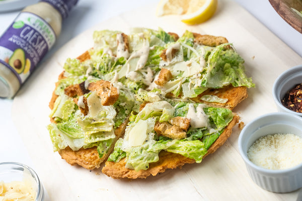 A Caesar salad pizza made with a canned chicken crust, next to a bottle of Primal Kitchen Caesar Dressing with a purple label.