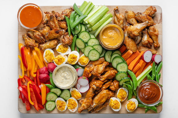 Overhead view of a charcuterie board with wings, veggies, and deviled eggs, with bowls of Primal Kitchen sauces and dips.