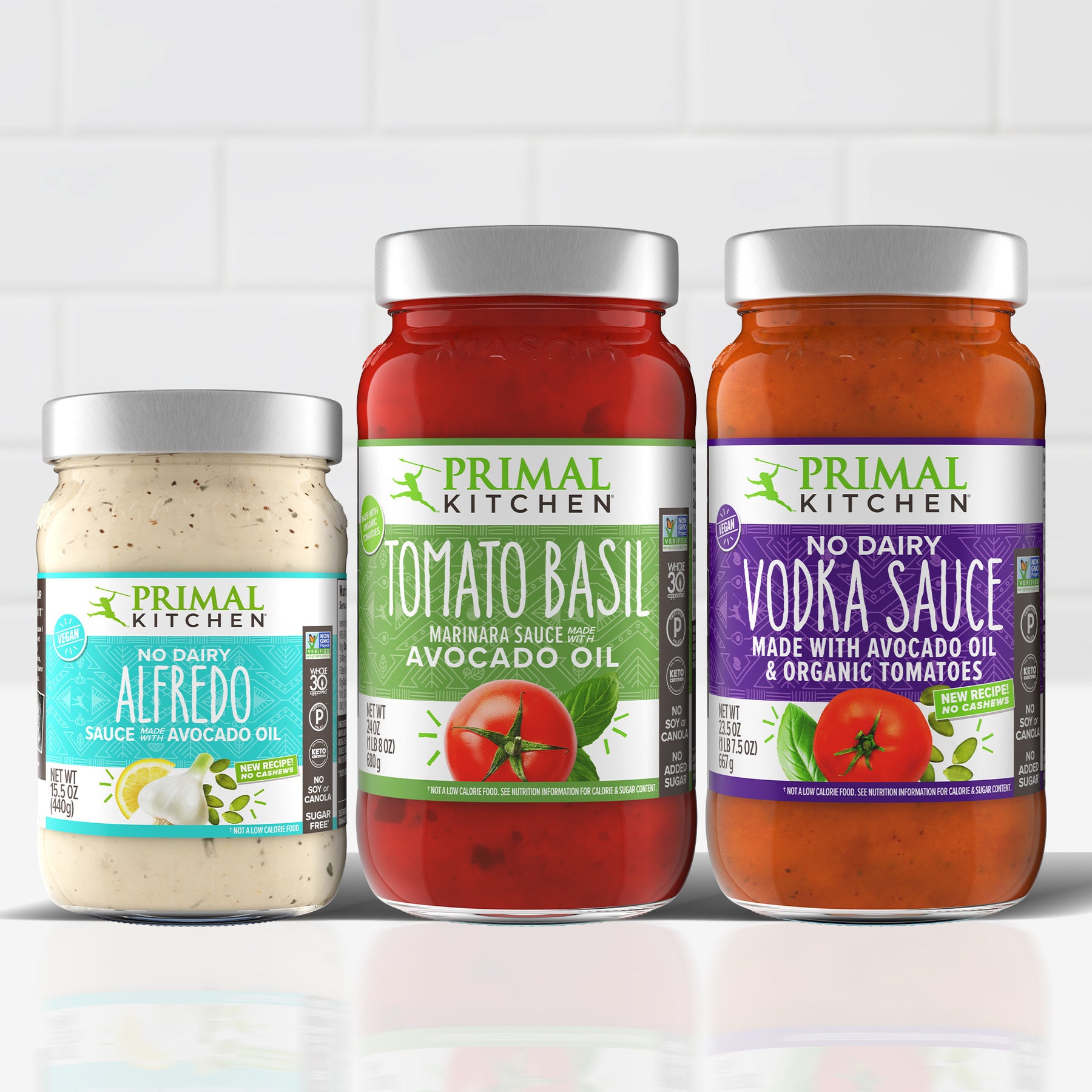 Jars of Primal Kitchen No Dairy Alfredo Sauce, Primal Kitchen Tomato Basil Marinara Sauce, and Primal Kitchen No Dairy Vodka Sauce Made with Avocado Oil and Organic Tomatoes lined up on a white countertop, with a white tile background. 