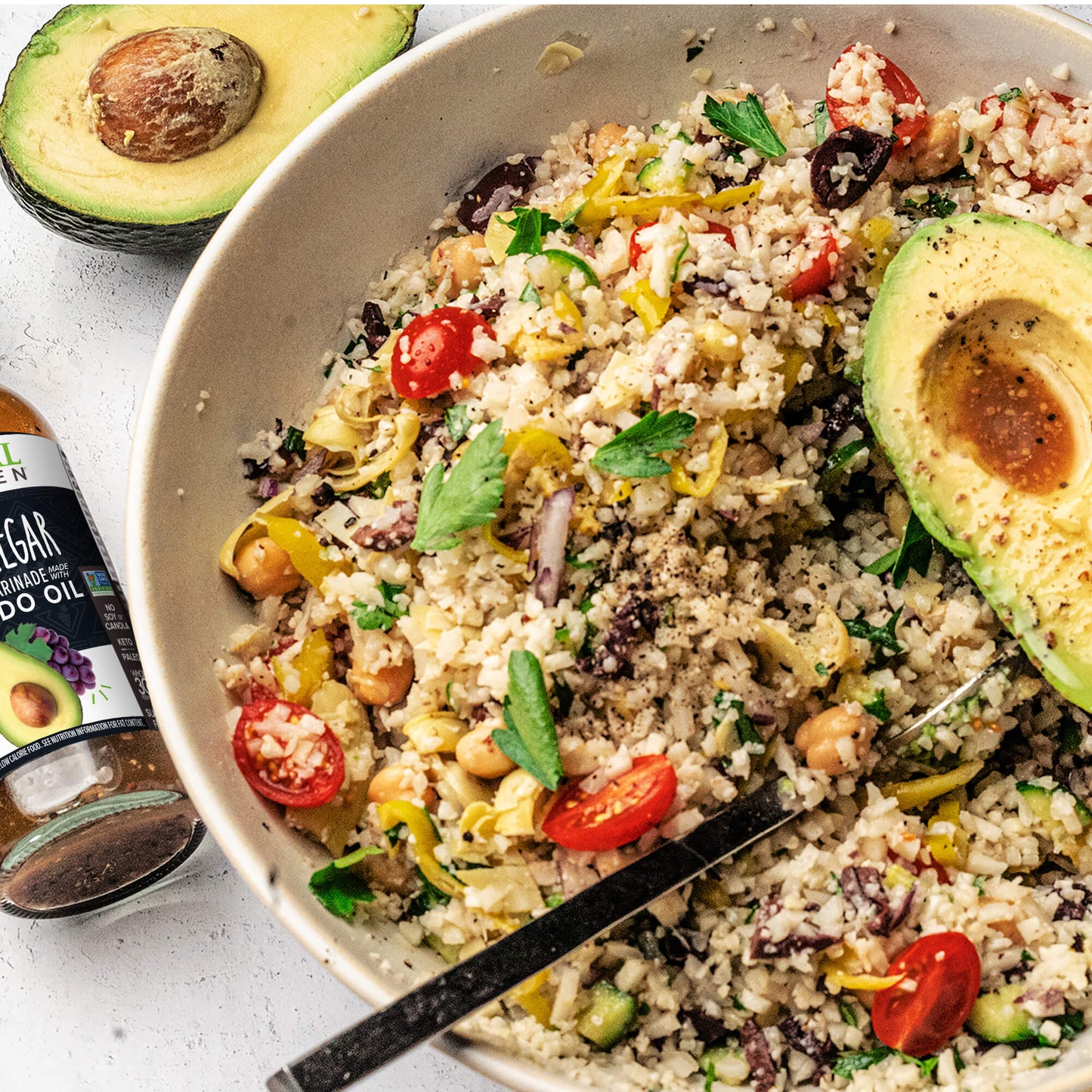 A veggie grain bowl with avocado and chickpeas, next to a bottle of Primal Kitchen Avocado Oil and Vinegar Dressing and a sliced avocado half.