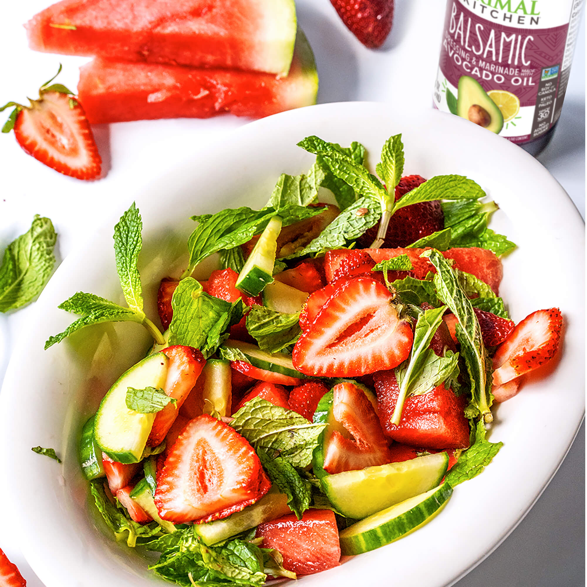 A mouthwatering cucumber mint and strawberry salad in a white dish, with watermelon slices and a bottle of Primal Kitchen Balsamic Dressing and Marinade in the background. 