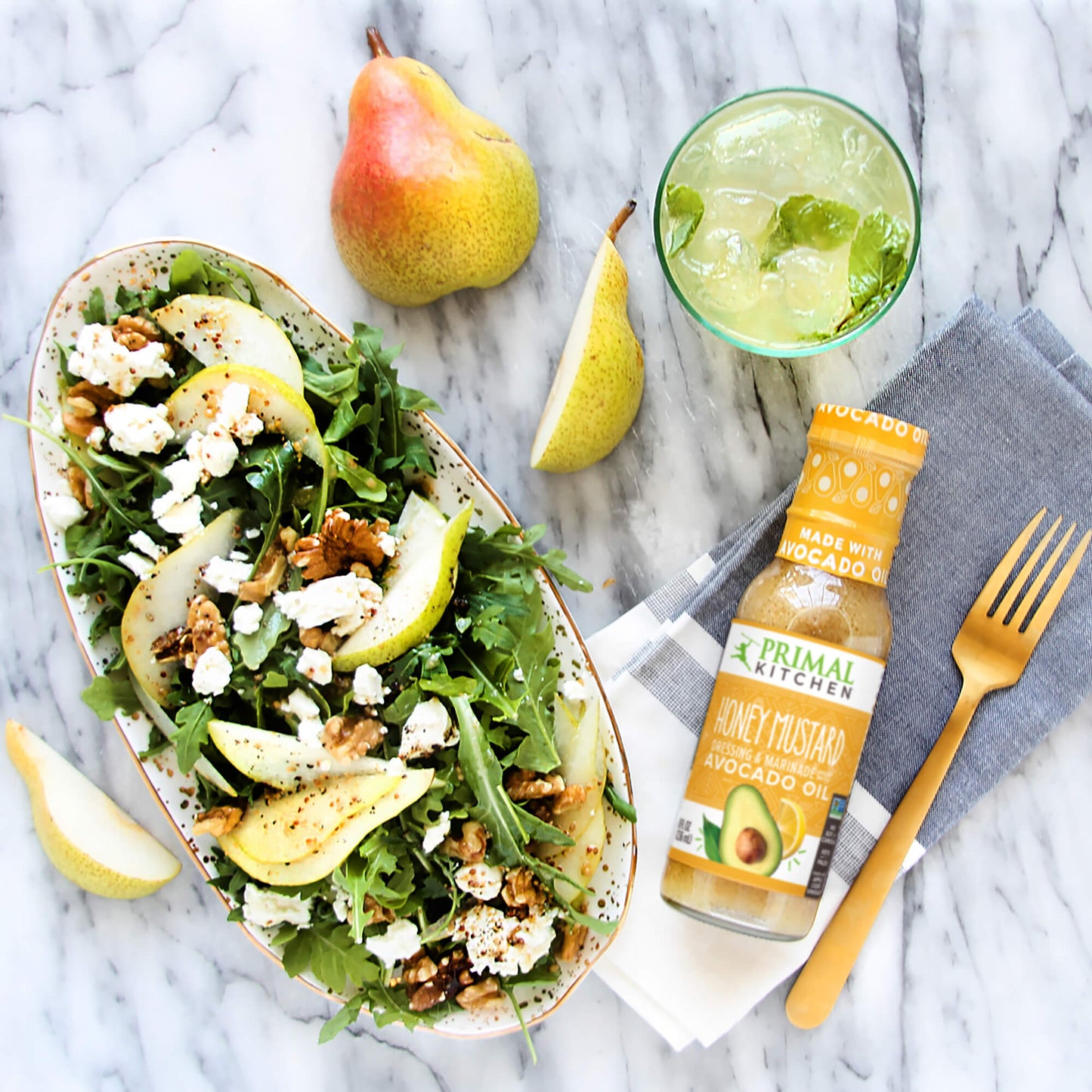 A pear and walnut salad with crumbled feta in a bowl, next to a bottle of Primal Kitchen Honey Mustard Dressing. Napkin, fork, drink, and sliced pear in the background.