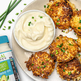 Vegan crab cakes sprinkled with chives on a white plate with a cup of Vegan Mayo next to cut chives and a jar of Primal Kitchen Vegan Mayo.