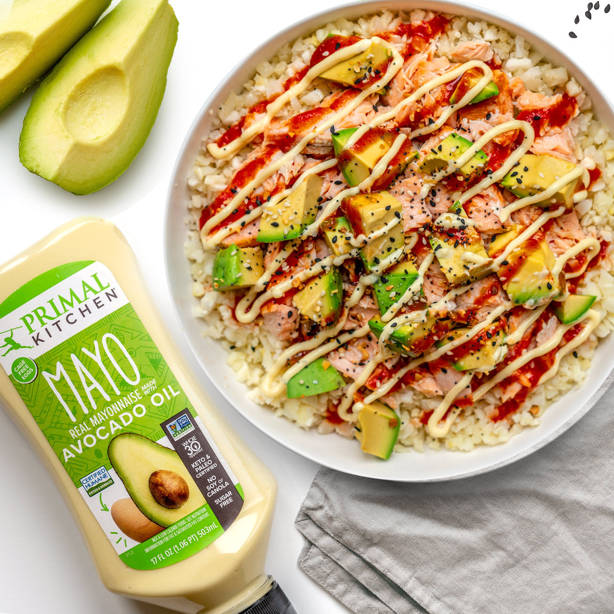 Avocado salmon rice bowl drizzled with mayo in a white bowl on top of a gray napkin next to cut avocado and a squeeze bottle of Primal Kitchen Mayo.