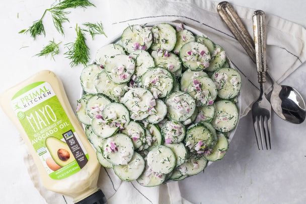 A bowl of creamy cucumber salad on a white table cloth next to a bottle of Primal Kitchen Squeeze Mayo