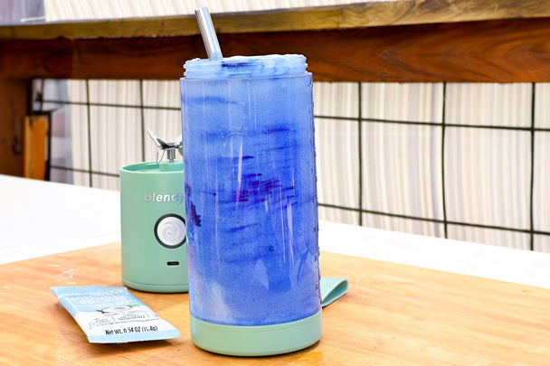 A bright blue Coconut Cloud Smoothie in a BlendJet, with a metal straw in it, displayed on a wooden table.