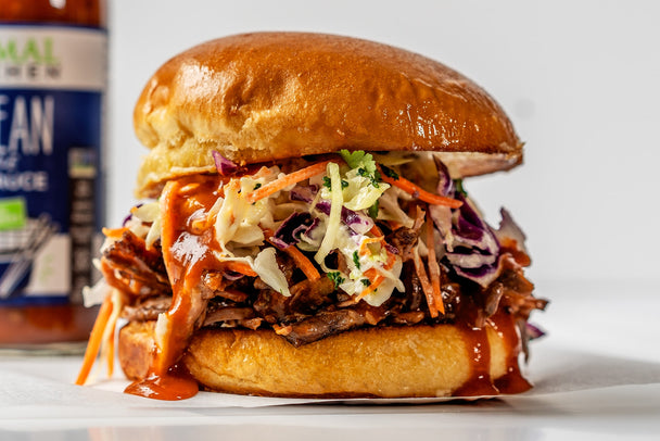 A short rib sandwich topped with coleslaw, on a toasted bun. In the background, a bottle of Primal Kitchen Korean Style BBQ Sauce.