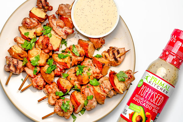 Five chicken and apple kabobs on a white plate with a small ramekin of dressing, next to a bottle of Primal Kitchen Poppyseed Dressing. 