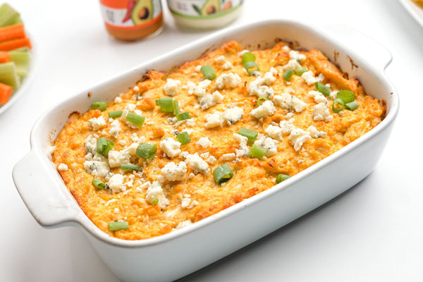 A white baking dish filled with Easy Buffalo Chicken Dip made with Primal Kitchen Original Buffalo Sauce. Chips, pretzels, and celery and carrot sticks in the background.