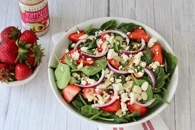  A white bowl of strawberry spinach salad with a bottle of Primal Kitchen Poppyseed Dressing, a bowl of strawberries, and a napkin in the background. 