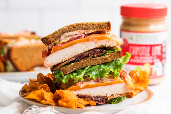 Two stacked halves of a Chicken Chipotle Sandwich made with Primal Kitchen Chipotle Lime Mayo, on a white plate with a side of chips. A jar of mayo, napkins, and another sandwich in the background.  