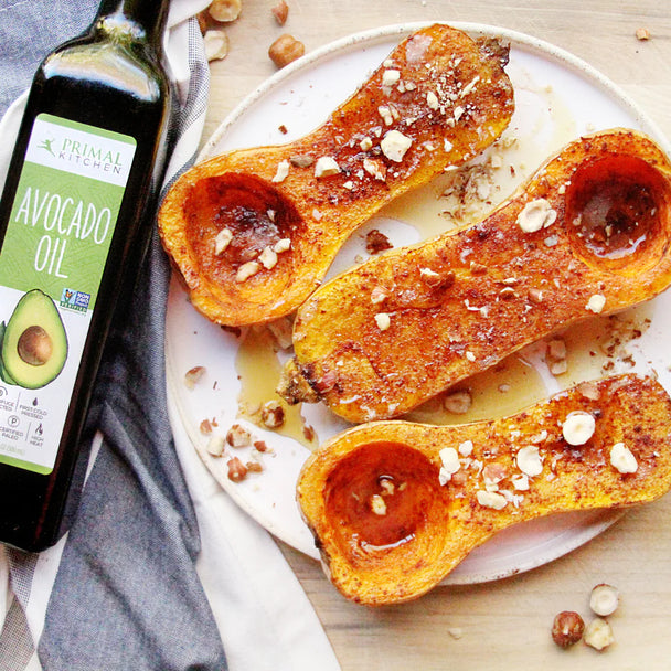 Three roasted butternut squash halves drizzled with maple syrup, on a plate next to a bottle of Primal Kitchen Avocado Oil.