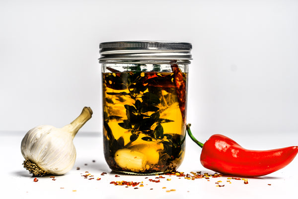 A mason jar of garlic chili infused avocado oil made with Primal Kitchen Avocado Oil next to a red Fresno pepper and a garlic bulb.