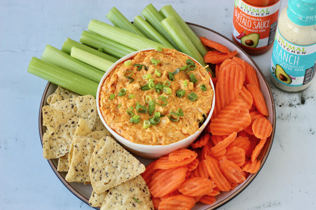 A round platter of vegetables and crackers with crock pot Buffalo dip in the center. Bottles of Primal Kitchen Ranch and Buffalo Sauce in the background.