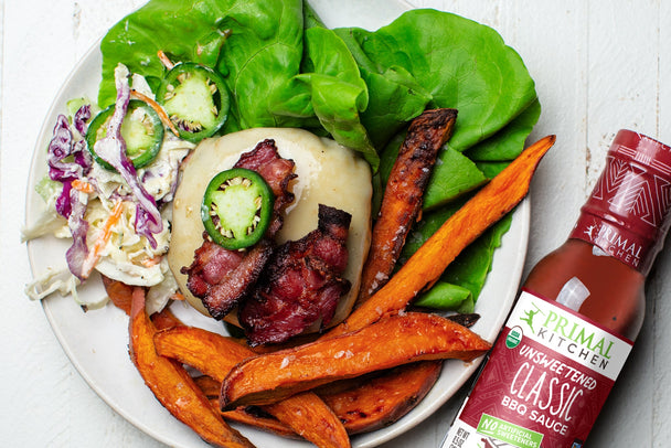 A spicy bbq with no bun on a white plate with sweet potato fries and lettuce, next to a bottle of Primal Kitchen BBQ Sauce. 
