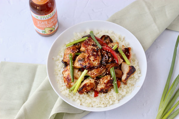 Sesame Ginger Chicken over riced cauliflower in a white dish, with a bottle of dressing on the side. 