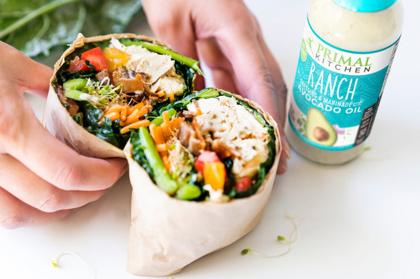 A hand holding a chicken bacon ranch collard green wrap split into halves, with a bottle of ranch dressing in the background.