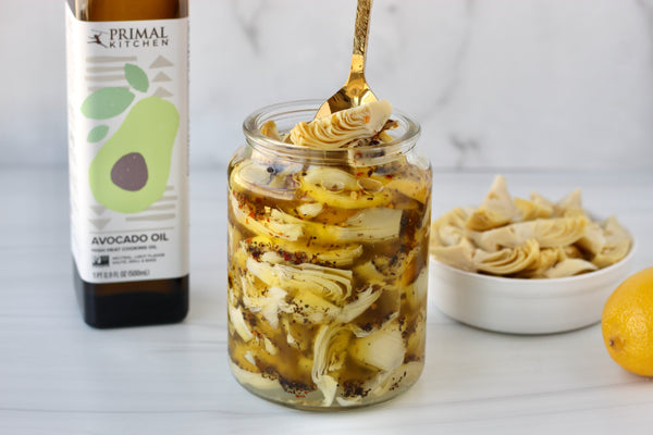 A jar of marinated artichoke hearts next to a bottle of Primal Kitchen Avocado Oil and a bowl of artichoke hearts. 