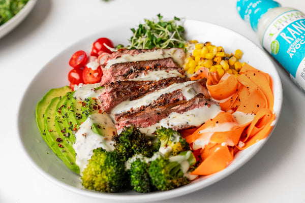  A protein bowl topped with steak, avocado, and veggies, next to a bottle of Primal Kitchen Ranch. 