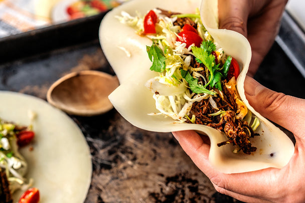 Hands hold a shredded beef taco in a cheese wrap with cabbage and tomatoes.