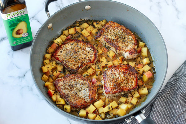 Pork chops and apples in a skillet, with a bottle of Primal Kitchen Avocado Oil in the background. 