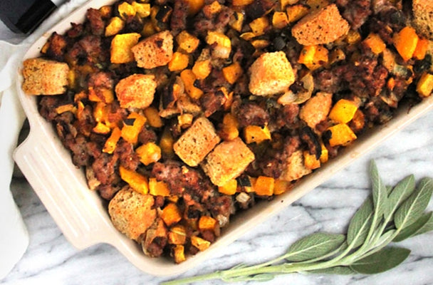 A white baking dish filled with stuffing, next to a bottle of Primal Kitchen Avocado Oil, a linen napkin, and a fresh herb.