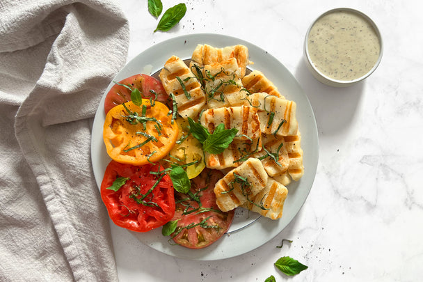 Grilled halloumi on white plate with sliced tomatoes (serving suggestion) and a ramekin of dipping sauce.