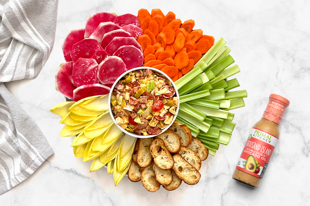 Round platter of fresh vegetables surrounding a bowl of hoagie dip with a bottle of Primal Kitchen Thousand Island Dressing.