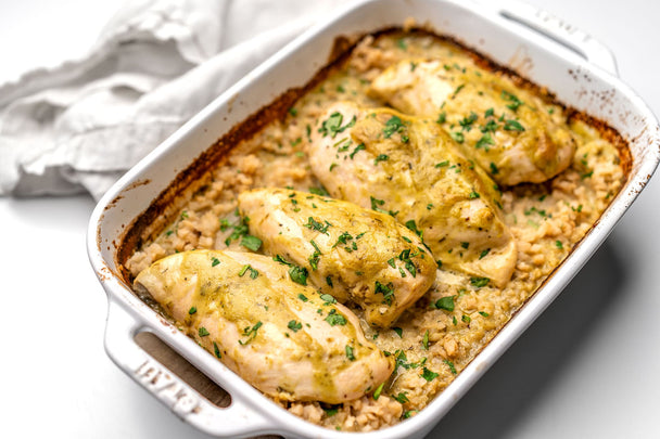 Four baked chicken breasts with Primal Kitchen Avocado-Lime Sauce & Dressing, in a white ceramic baking dish next to a white linen napkin. 