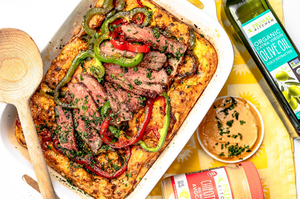 Steak fajita frittata in a white ceramic dish, with a wooden spoon on top, next to a ramekin of Chipotle Lime Mayo and a bottle of Primal Kitchen olive oil.