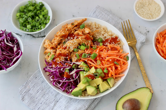 A large bowl of cauliflower rice, shredded chicken, and colorful veggies made with Primal Kitchen Korean Style BBQ Sauce, with an avocado half and small bowls of fresh veggies in the background. 