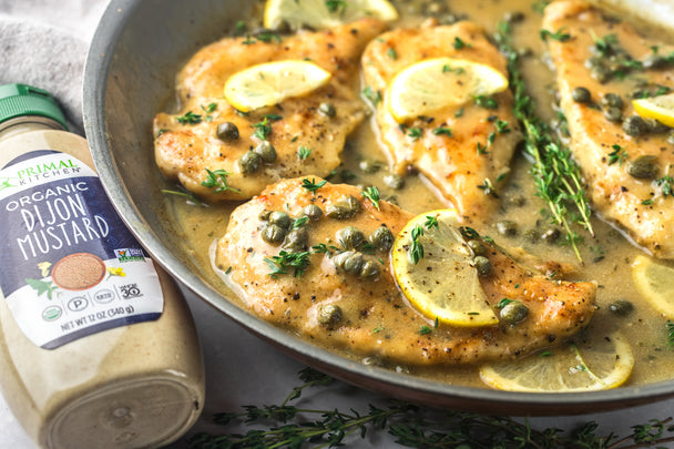 Lemon chicken picatta in a skillet, next to a bottle of Primal Kitchen Dijon Mustard.