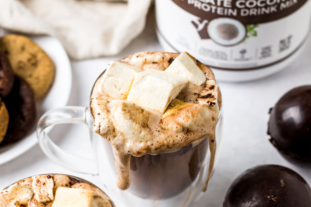 Close up of clear mug with a melted hot chocolate bomb oozing down the sides of the mug. Hot chocolate is topped with marshmallow cubes. Primal Kitchen Primal Fuel is in the background, along with a plate of cookies and hot chocolate bombs.