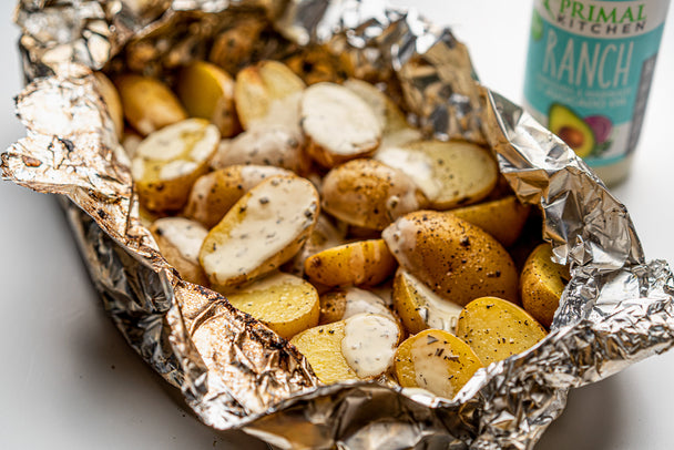 Grilled potatoes in foil are covered in ranch dressing with the bottle behind them.