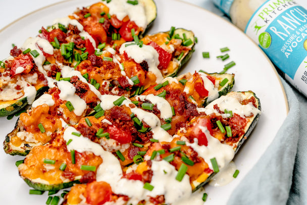 A white plate of cheesy zucchini boats topped with a drizzle of ranch, next to a bottle of Primal Kitchen Ranch Dressing.