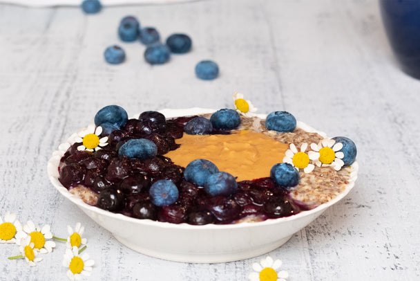Noatmeal in a large white bowl topped with stewed blueberries, fresh blueberries, almond butter, and baby daisy flowers. Fresh blueberries and daisies scattered around the bowl.