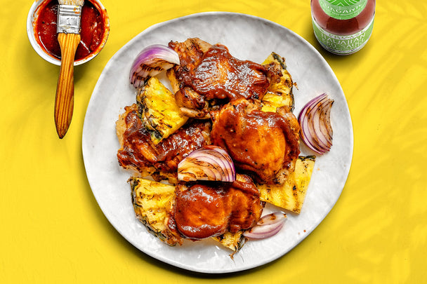 A white plate of Hawaiian BBQ Chicken on a yellow background next to a bottle of Primal Kitchen Hawaiian Style BBQ Sauce and a small bowl of sauce with a basting brush.
