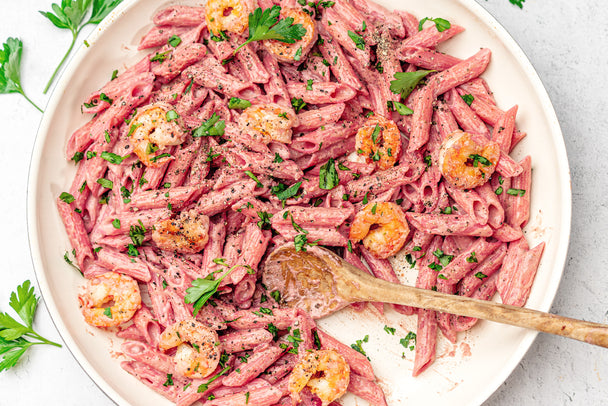 A large white bowl filled with penne with pink sauce and shrimp, topped with chopped parsley. A wooden spatula is in the bowl, with a little bit of penne missing. Primal Kitchen Pink Primavera Sauce is next to the bowl.
