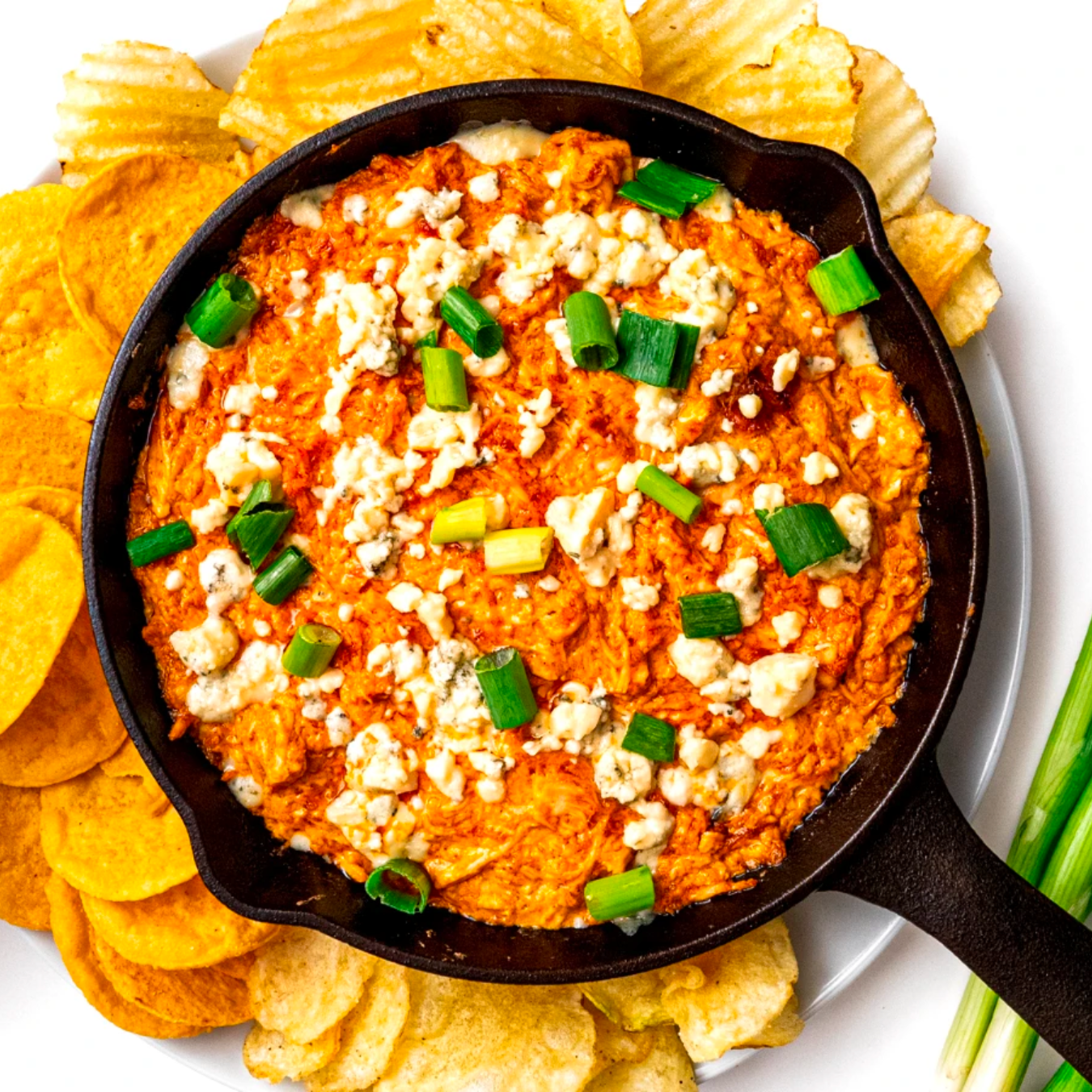 Buffalo Chicken Dip in a cast iron skillet, topped with green onions & blue cheese, with a side of potato chips.