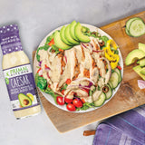 A mouthwatering chicken Caesar salad with creamy Caesar salad dressing  and veggies on a cutting board , next to a bottle of Primal Kitchen No Dairy Caesar Dressing & Marinade. 
