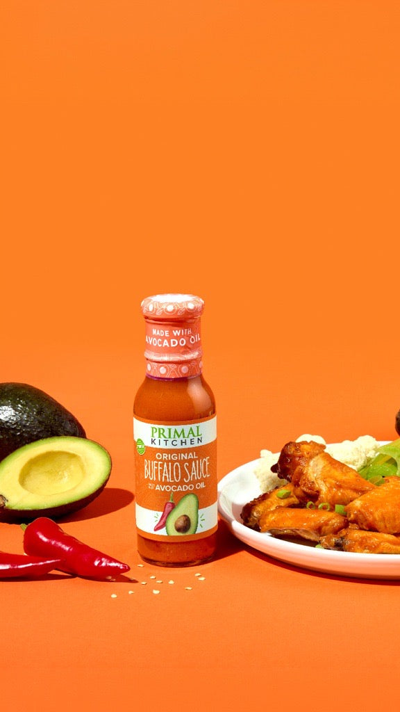 A bottle of Primal Recipe Buffalo Sauce surrounded by food props on a bright orange background, including a plate of buffalo wings with cauliflower and celery, avocado halves, red chili peppers, and a small dipping cup.