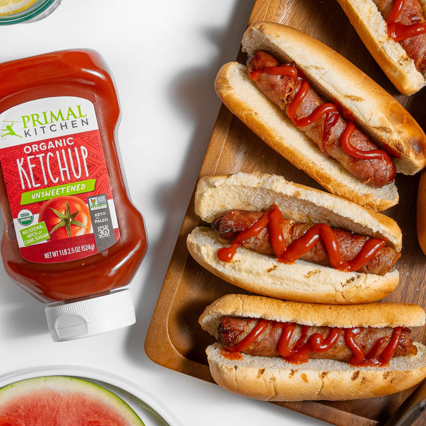 Primal Kitchen ketchup bottle with hot dogs on buns topped with ketchup on a wooden board.