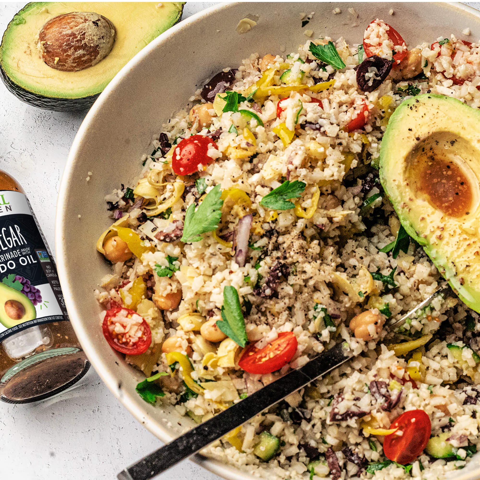 A veggie grain bowl with avocado  and chickpeas, next to a bottle of Primal Kitchen Avocado Oil and Vinegar Dressing and a sliced avocado half.