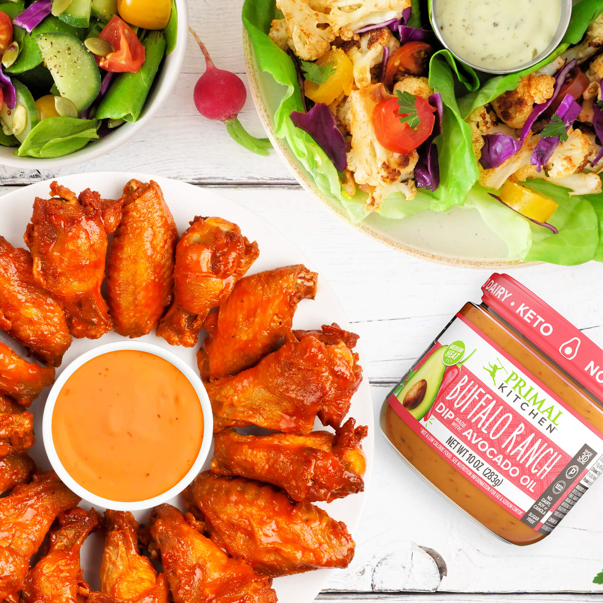A garden salad, a plate of lettuce cups, and a white plate of Buffalo chicken wings next to a jar of Primal Kitchen Buffalo Ranch Dip. 