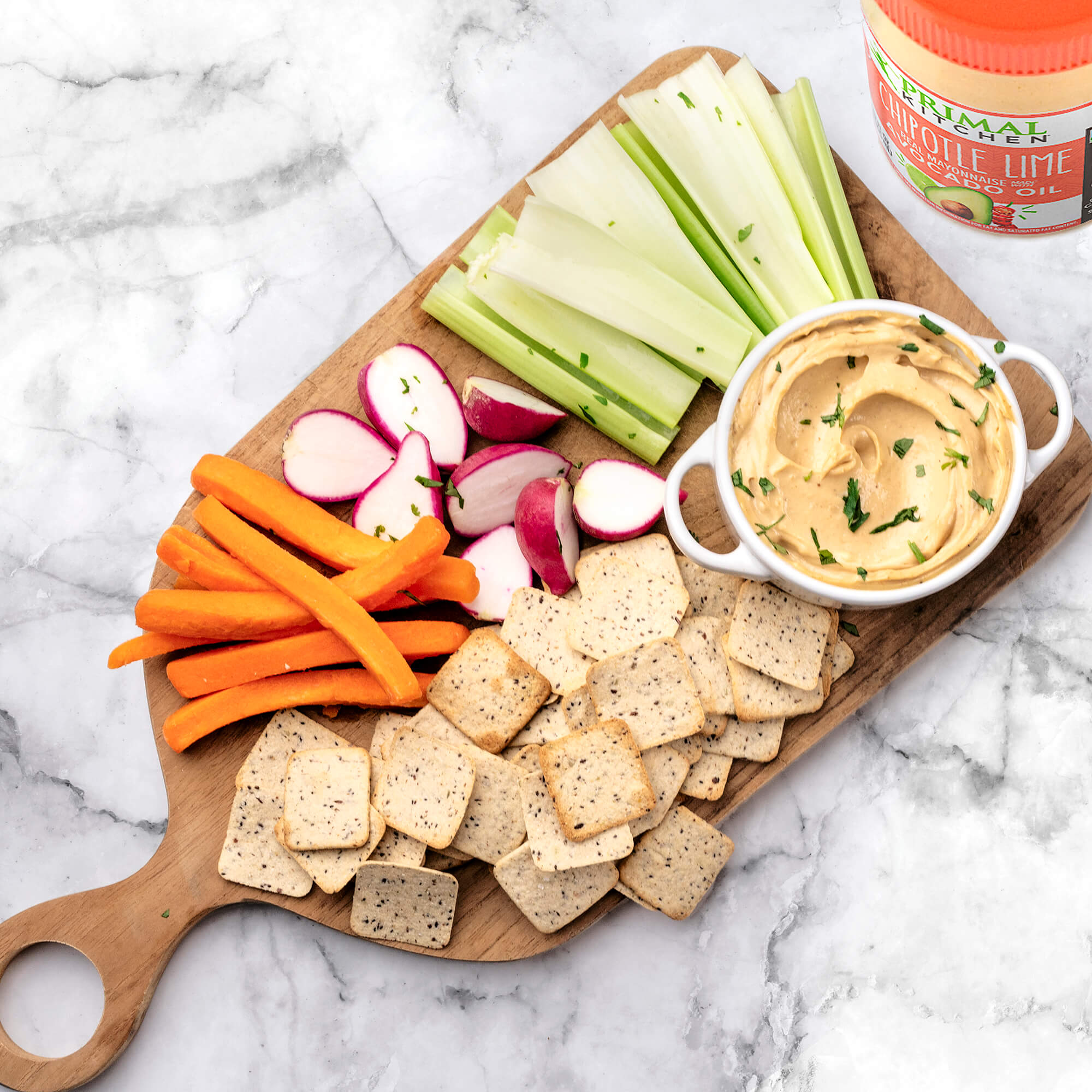 Snack spread of carrots, crackers, radishes, celery and Primal Kitchen Chipotle Lime Mayo in a white car on a wooden board next to a jar of Primal Kitchen Chipotle Lime Mayo.